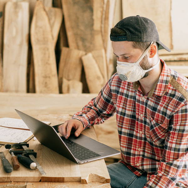 Handwerksbetrieb Schreinerei. Der Meister mit Schutzmaske sitzt an der Werkbank und schaut auf seinen Laptop-Computer.