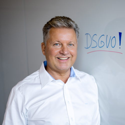 Mannus Weiß der Geschäftsführer der Datenschutzkonzept GmbH in einem weißen Hemd vor einer Tafel auf der DSGVO steht.
