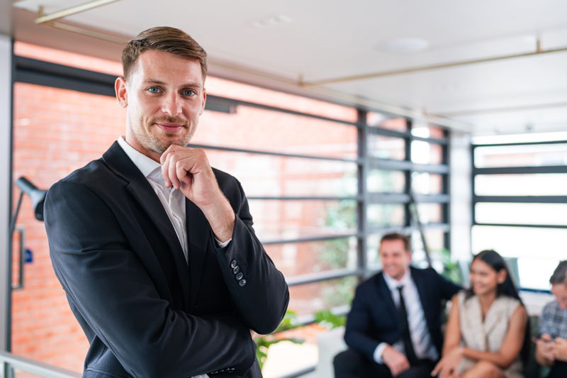 Ein junger Mann im dunklen Anzug in steht in einem sehr hellen Büro vor einer Gruppe sitzenden Menschen.