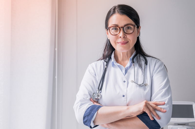 Eine Ärztin mit dunklen Haar und Brille, die ein Stethoskop um den Hals trägt.