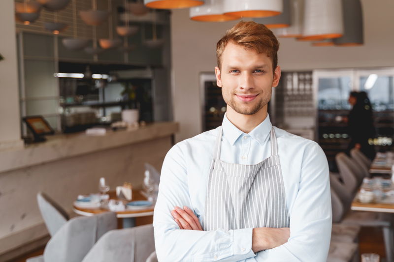 Ein junger Geschäftsführer mit Servirschürze steht mit verschränkten Armen in einem Restaurant.