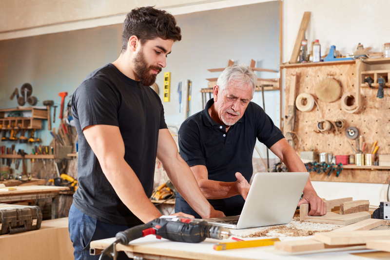 Ein Schreiner und sein Mitarbeiter schauen in der Werkstatt auf einen Laptop. Datenschutzfür alle Branchen, besonders für das Handwerk.