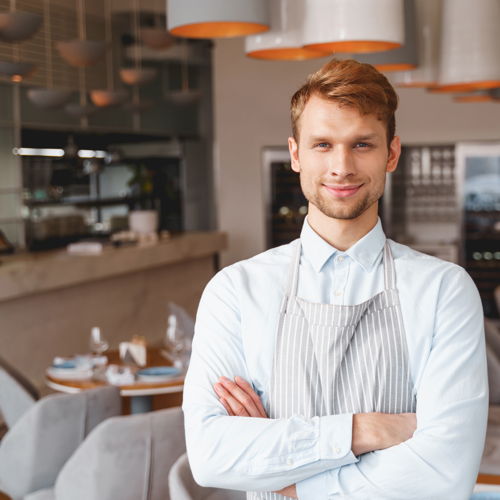 Ein junger Geschäftsführer mit Servirschürze steht mit verschränkten Armen in einem Restaurant.