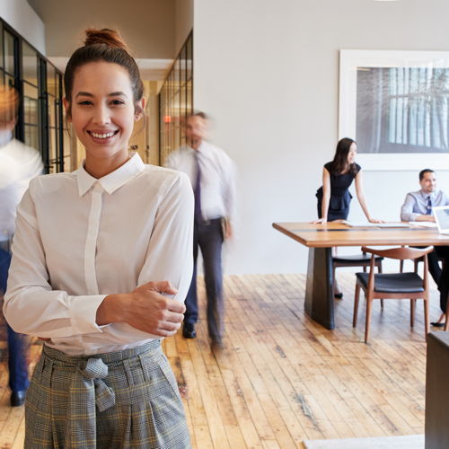 unternehmen Eine junge Frau in einem sehr modernen Büro. Hinter ihr befinden Kollegen, die am arbeiten sind.